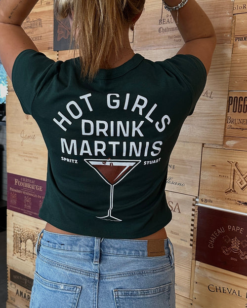 A woman viewed from behind wearing a dark green graphic t-shirt with the text 'HOT GIRLS DRINK MARTINIS' and a martini glass illustration, paired with light blue jeans, standing in front of a wall of wine crates.