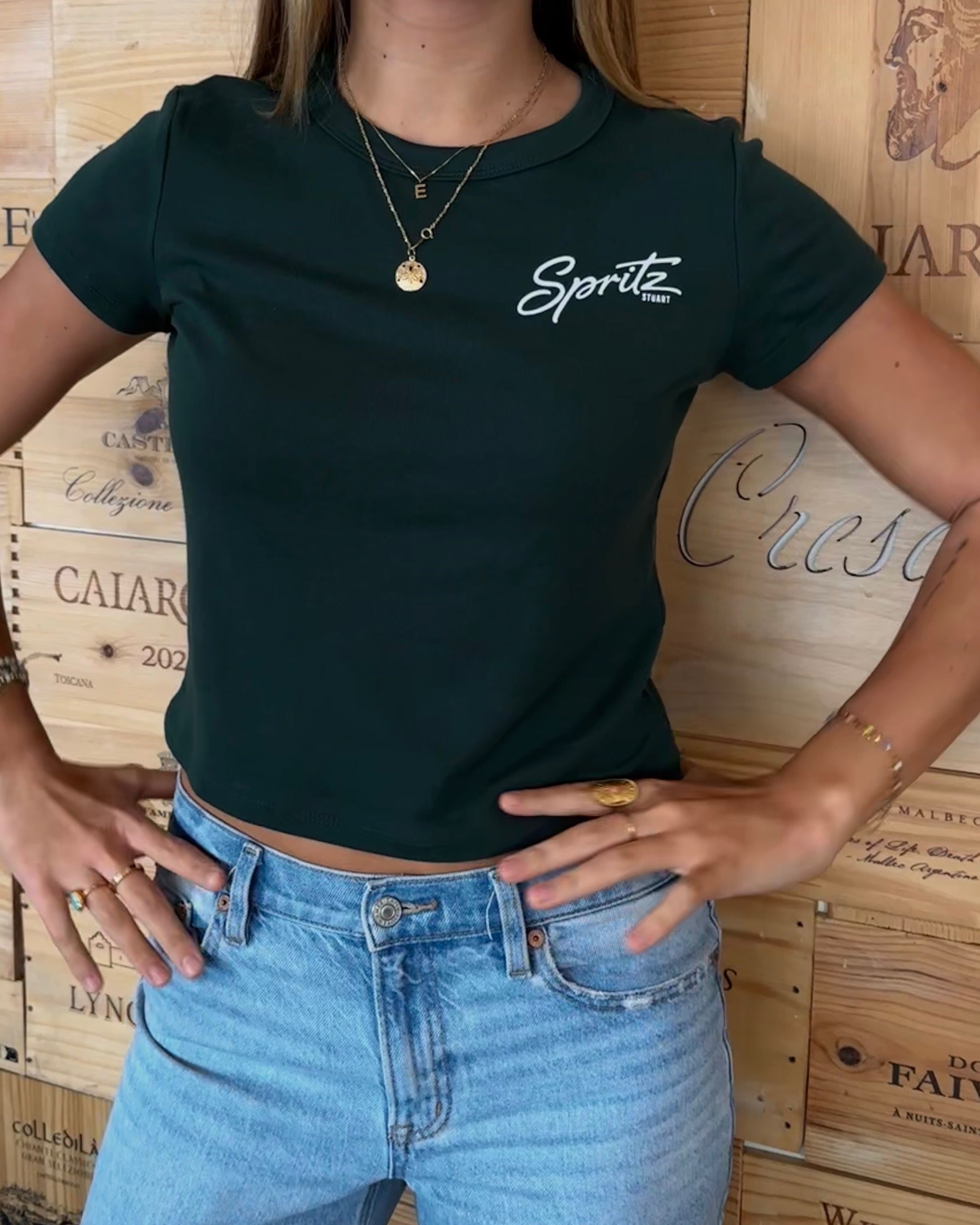 A woman wearing a dark forest green short-sleeve crop top with a white 'Spritez' script logo on the chest, paired with light blue jeans, standing in front of a wooden wine crate backdrop.