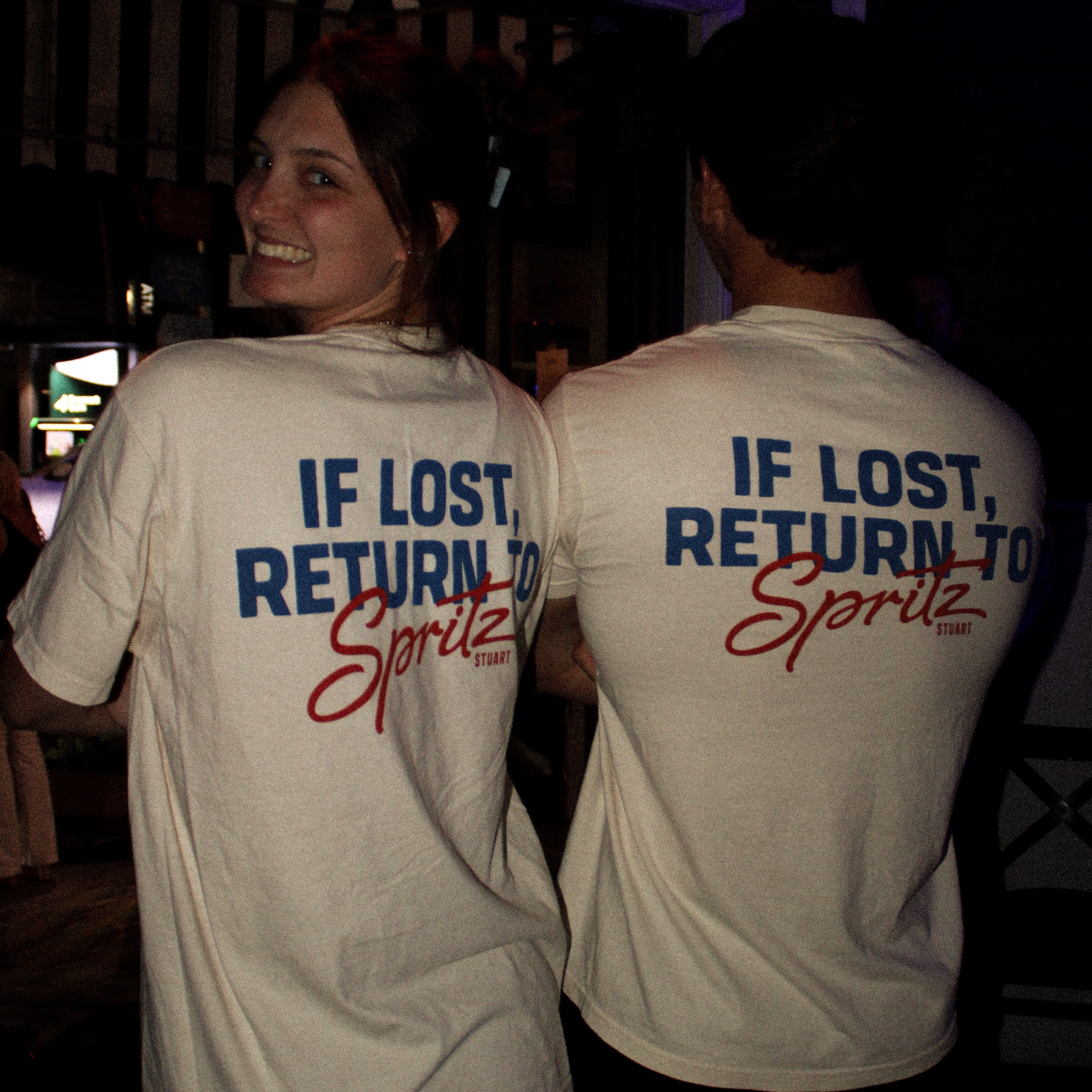 Two people wearing white t-shirts with text on the back, standing in a dark indoor setting.