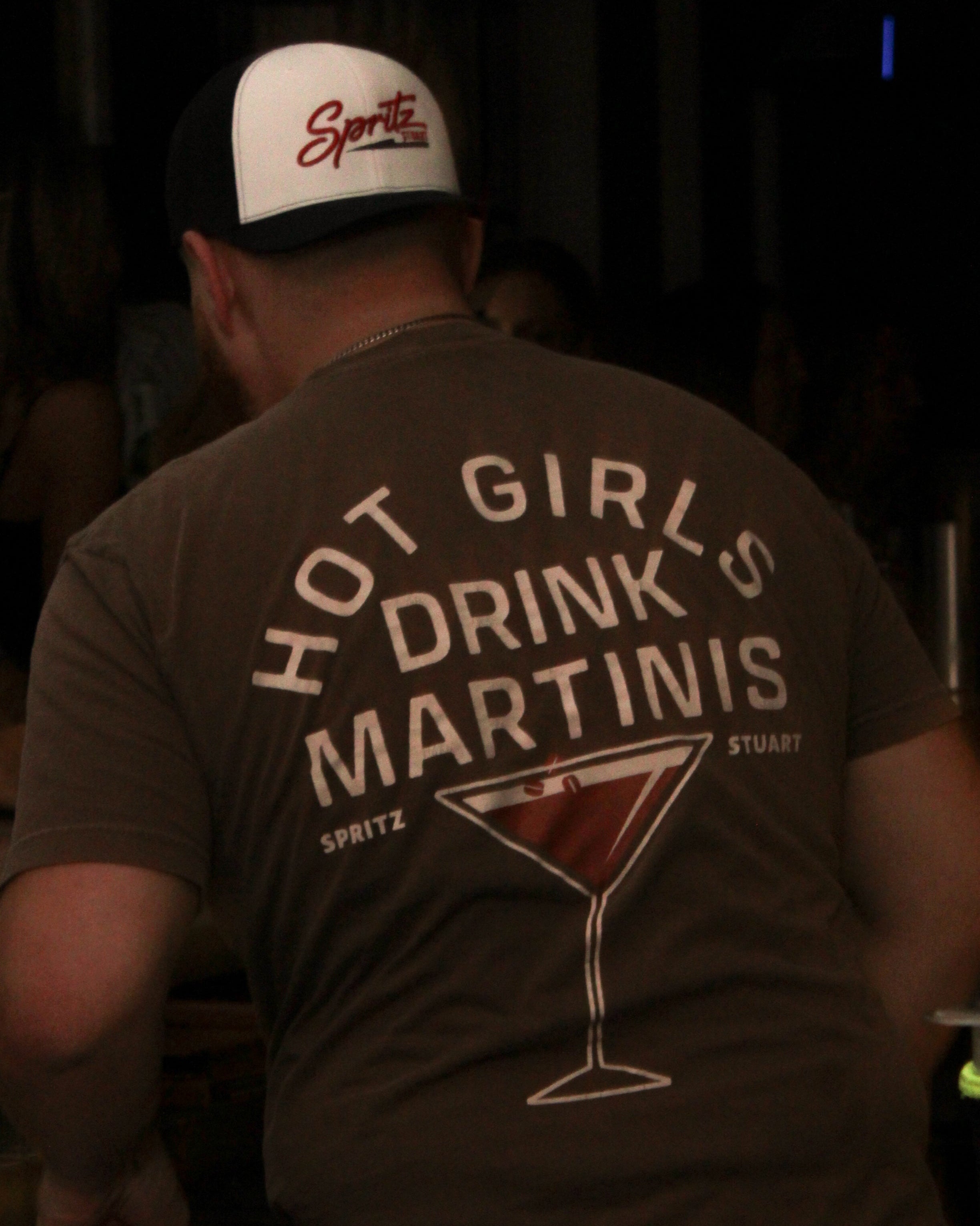 A man seen from behind wearing a brown t-shirt with a large white graphic on the back that reads 'HOT GIRLS DRINK MARTinis' and 'SPRITZ STUART' with a martini glass illustration. He is also wearing a white and black trucker hat with 'Spritz' written on it.