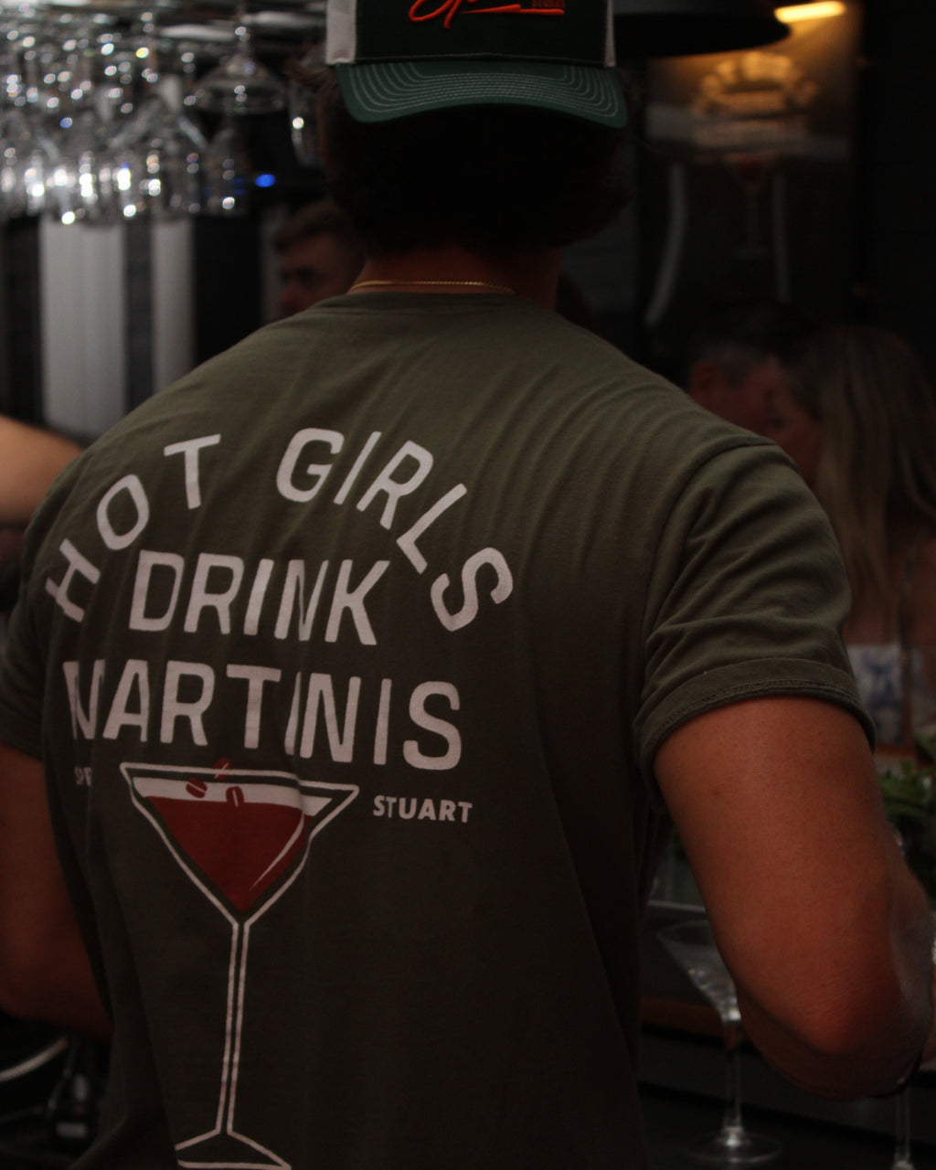 A person standing at a bar, shown from behind wearing an olive green t-shirt with a large white graphic on the back that reads 'HOT GIRLS DRINK MARTINIS' and features a martini glass illustration. The person is also wearing a dark green and white cap.
